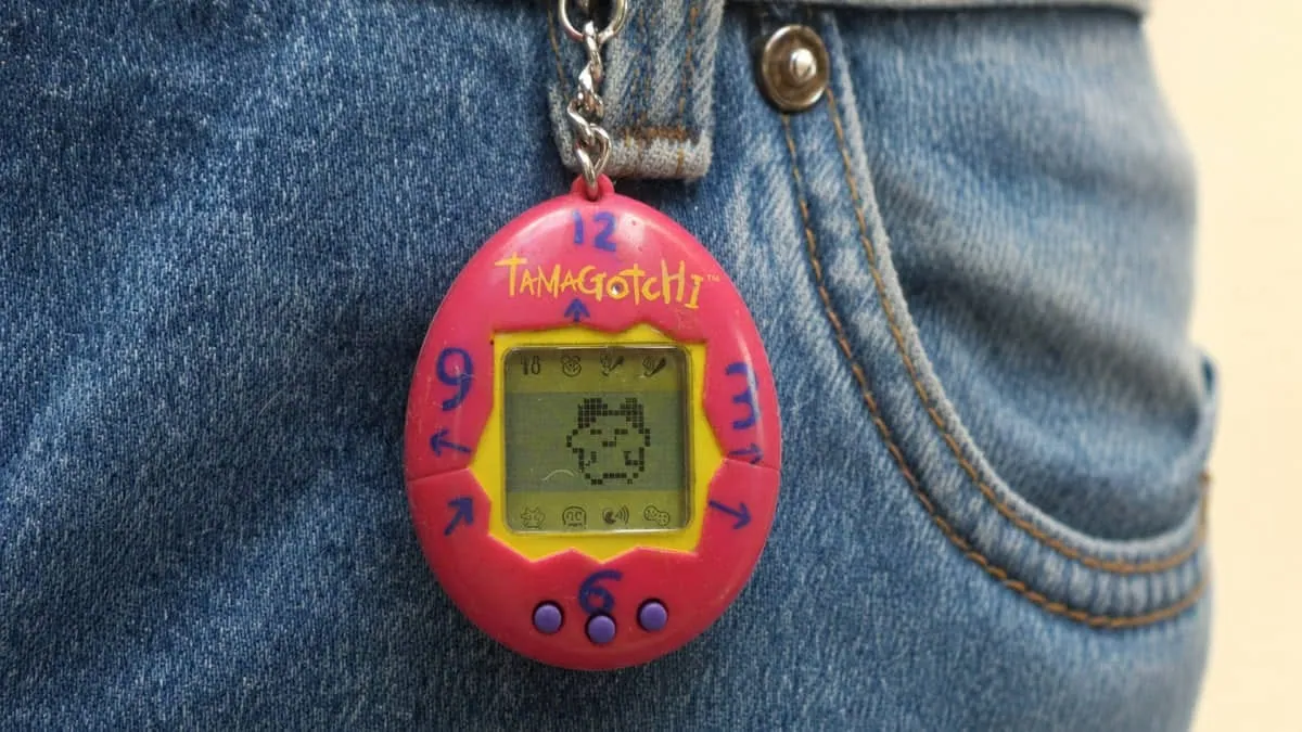 A pink 1990s Tamagotchi virtual pet clipped to the pocket of a pair of blue jeans, with a pixel creature visible on its screen.